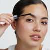Eine Person, die das transparente Gel XLASH Brow Booster mit einem maskarähnlichen Applikator auf ihre Augenbrauen aufträgt.