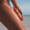 Am Strand trägt jemand das „Bali Body Sheer Glow Sunscreen Gel SPF 50“ auf seinen Oberschenkel auf, die orangefarbene Flasche in der Hand, das Meer im Hintergrund.