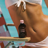 Eine Flasche Watermelon Sunscreen Oil SPF50 von Bali Body steht auf einer Sonnenstuhllehne, dahinter sitzt eine gebräunte Frau.
