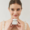 Eine Frau lächelt und hält einen Tiegel Ground Rice and Honey Glow Mask.