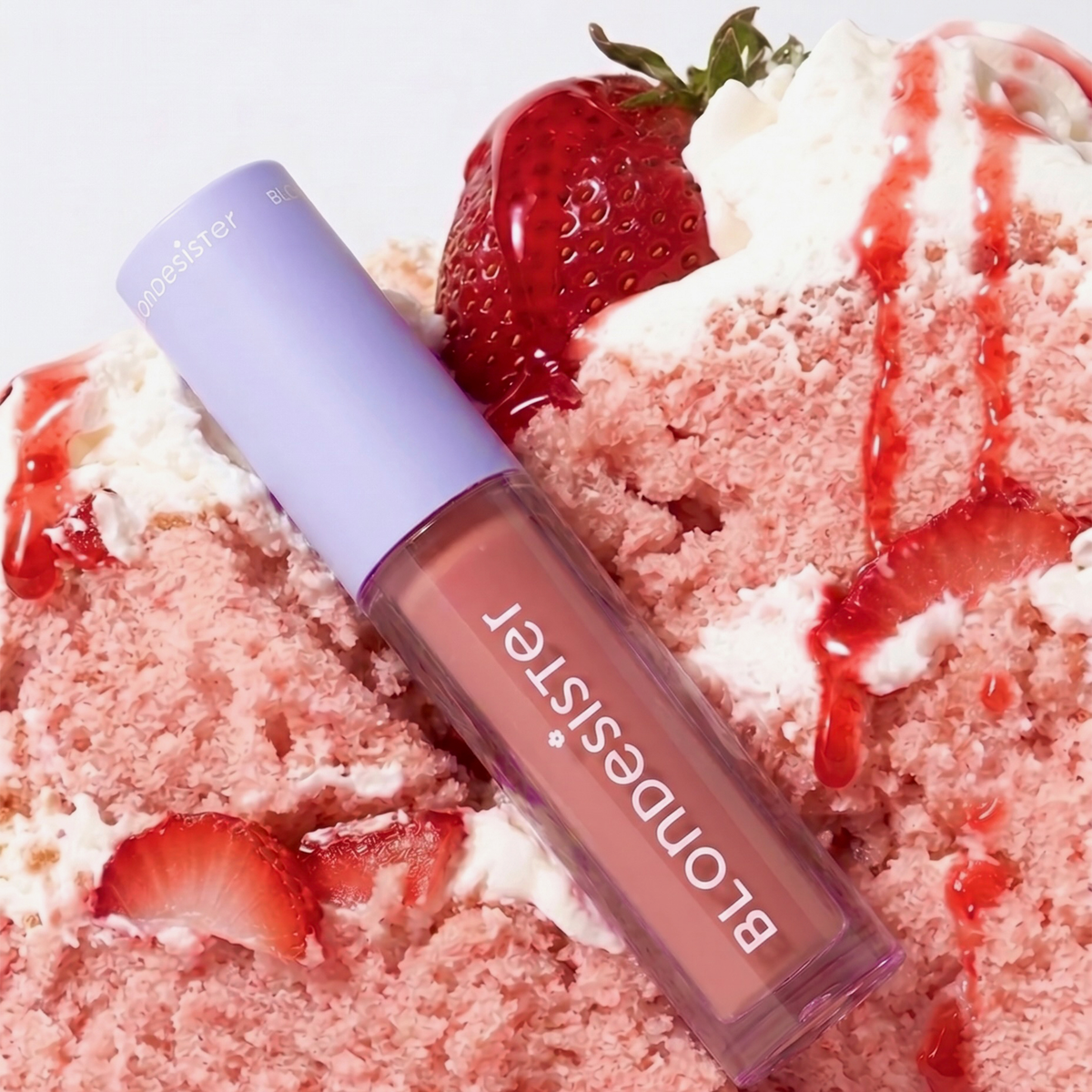 Eine Tube BLONDESISTER 2 IN 1 SWEET GLOSS TINTED LIP OIL liegt auf einem Erdbeerkuchen mit Schlagsahne und Erdbeersauce.