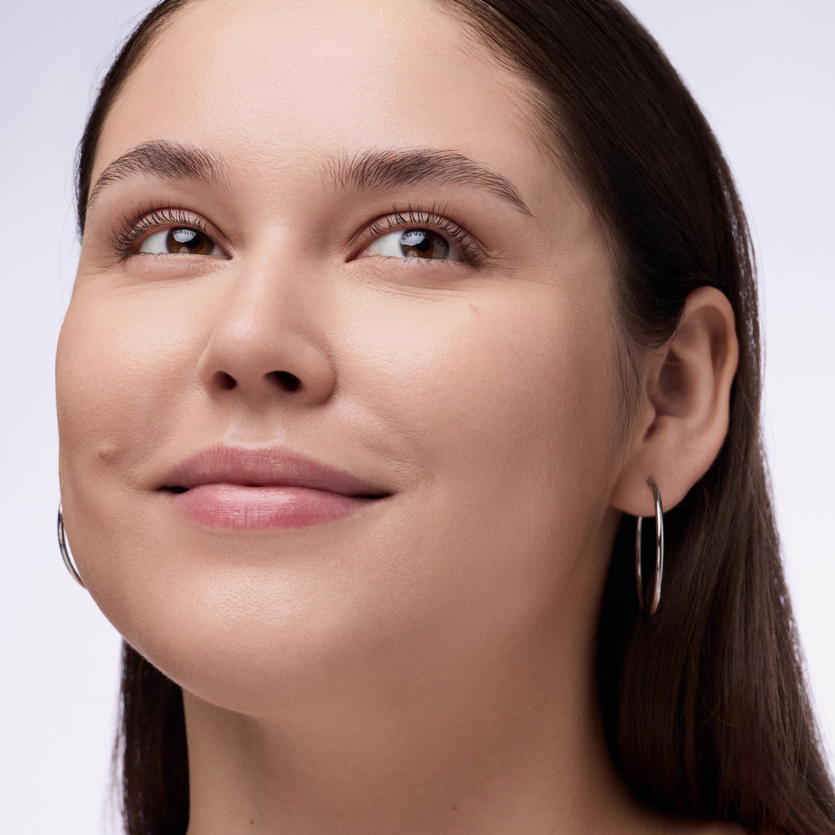 Nahaufnahme einer Person mit dunklem Haar und braunen Augen die den Instant Bright Serum Concealer 010C auf der Haut präsentiert.
