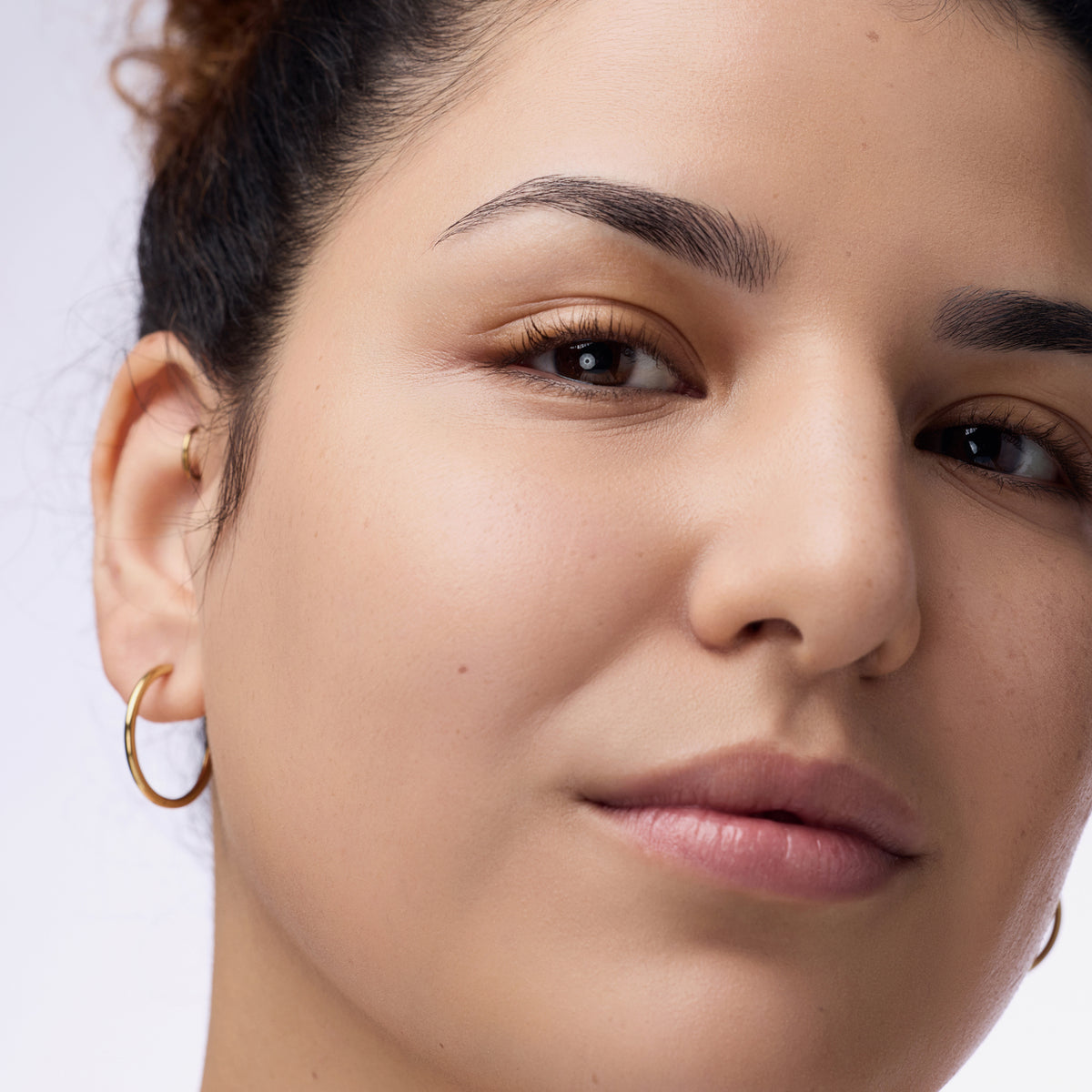 Nahaufnahme einer Person mit dunklem Haar und braunen Augen die den Instant Bright Serum Concealer 039W auf der Haut präsentiert.
