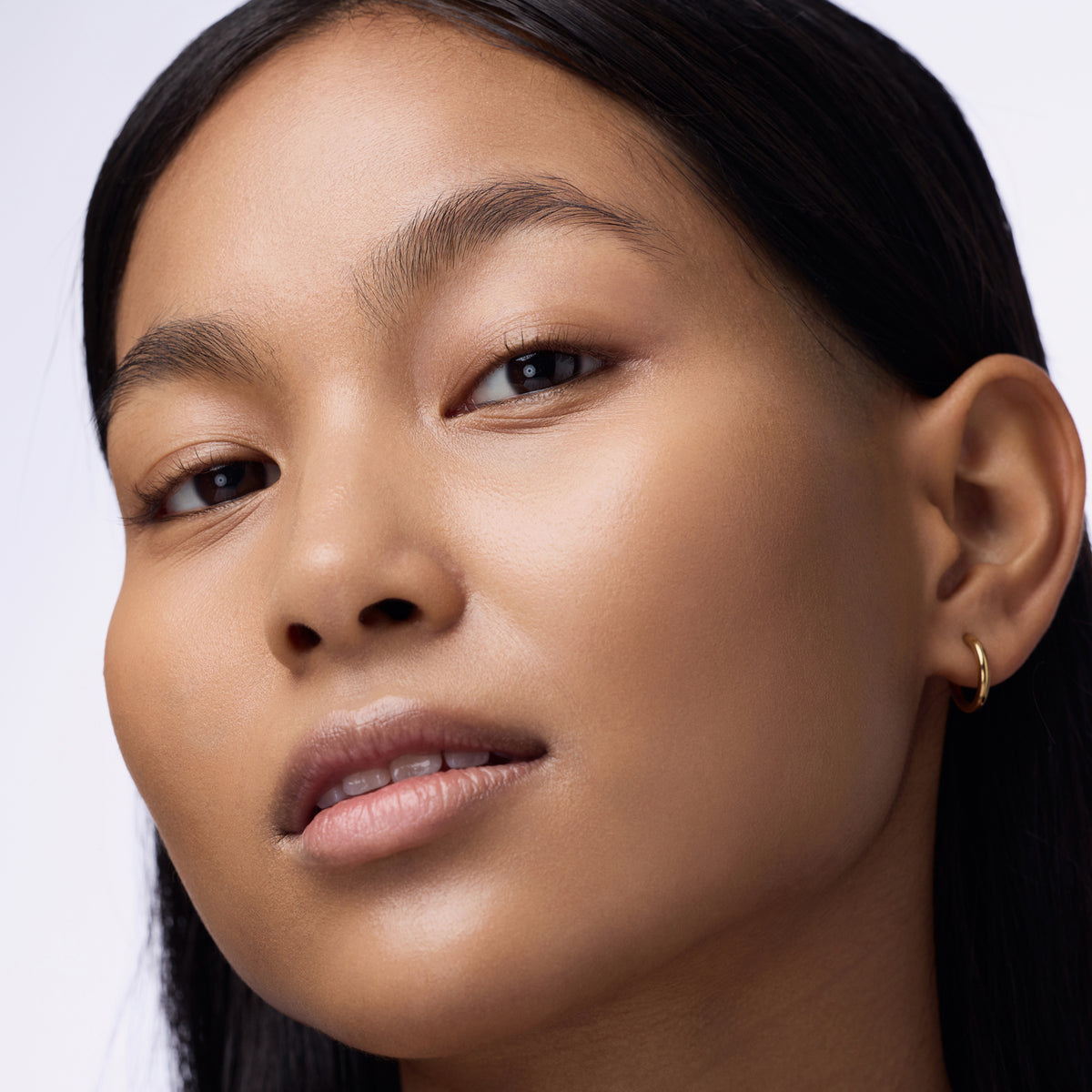 Nahaufnahme einer Person mit schwarzem Haar und braunen Augen die den Instant Bright Serum Concealer 060N auf der Haut präsentiert.
