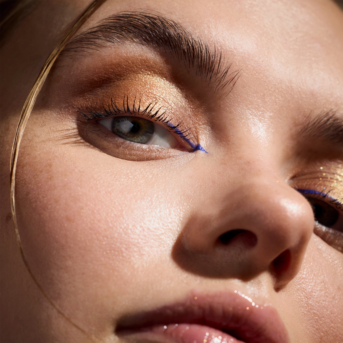 Nahaufnahme einer Person, die die Wonder Woman-Lidschattenpalette mit goldenen Farbtönen und blauem Eyeliner trägt und strahlende Haut und glänzende Lippen zur Schau stellt.