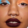 Eine Nahaufnahme zeigt eine Person mit blauem und orangefarbenem Colorfix Pastels-Lidschatten von Danessa Myricks Beauty, dunklem Lippenstift und mehreren Nasenpiercings.