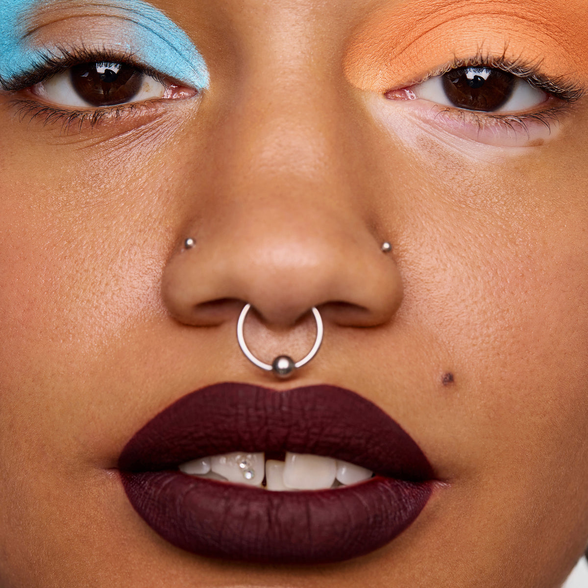 Eine Nahaufnahme zeigt eine Person mit blauem und orangefarbenem Colorfix Pastels-Lidschatten von Danessa Myricks Beauty, dunklem Lippenstift und mehreren Nasenpiercings.