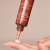 Eine Hand mit rosafarbenen Nägeln verteilt das Beauty Yummy Skin 10/10 Barrier Boost Serum aus einer braunen Tube auf die Fingerspitze vor einem beigen Hintergrund.