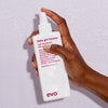 Eine Hand hält eine Flasche Baby Got Bounce Curl Treatment von Evo vor nassem Hintergrund.