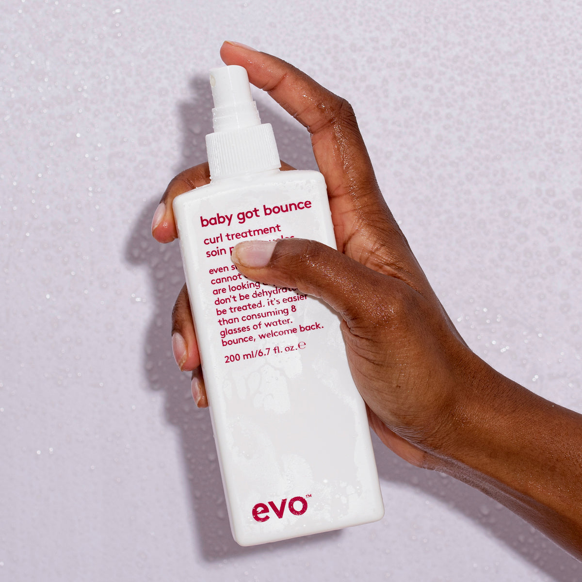 Eine Hand hält eine Flasche Baby Got Bounce Curl Treatment von Evo vor nassem Hintergrund.