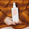 Eine Person in einem braun-orangem Pullover hält eine Flasche Happy Campers Wearable Treatment.