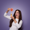 Frau mit langen dunklen Haaren hält eine Grapefruitscheibe über eine Hair-Syrup-Rapunzel-Flasche; Saft tropft hinein, lila Hintergrund.