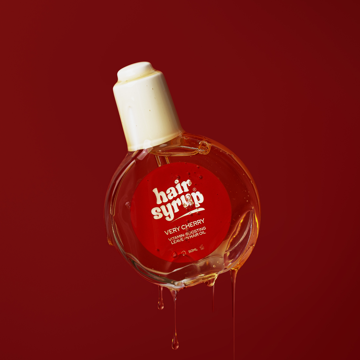 Eine Flasche Very Cherry von Hair Syrup schwebt for rotem Hintergrund, Haaröl läuft an der Falsche hinab.