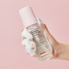 Eine Hand hält eine schaumige Flasche Real Complexion Hyaluron Bubble Pop Cleanser vor einem rosa Hintergrund.