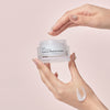 Eine Hand hält einen Tiegel mit der Real Complexion Hyaluron Moisture Cream,vor einem rosa Hintergrund.