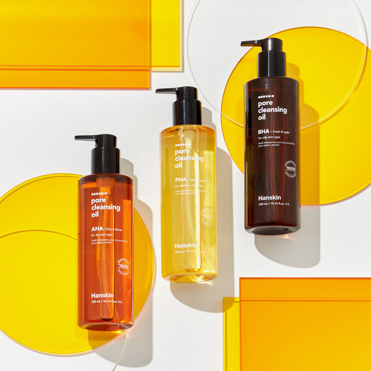 Drei Flaschen Pore Cleansing Oil AHA auf einer weißen Oberfläche mit gelben und orangefarbenen Glaskreisen präsentiert.