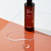 Eine Flasche Pore Cleansing Oil BHA mit auf einer orange-weißen Glasfläche; davor ein Tropfen des Reinigungsöls.