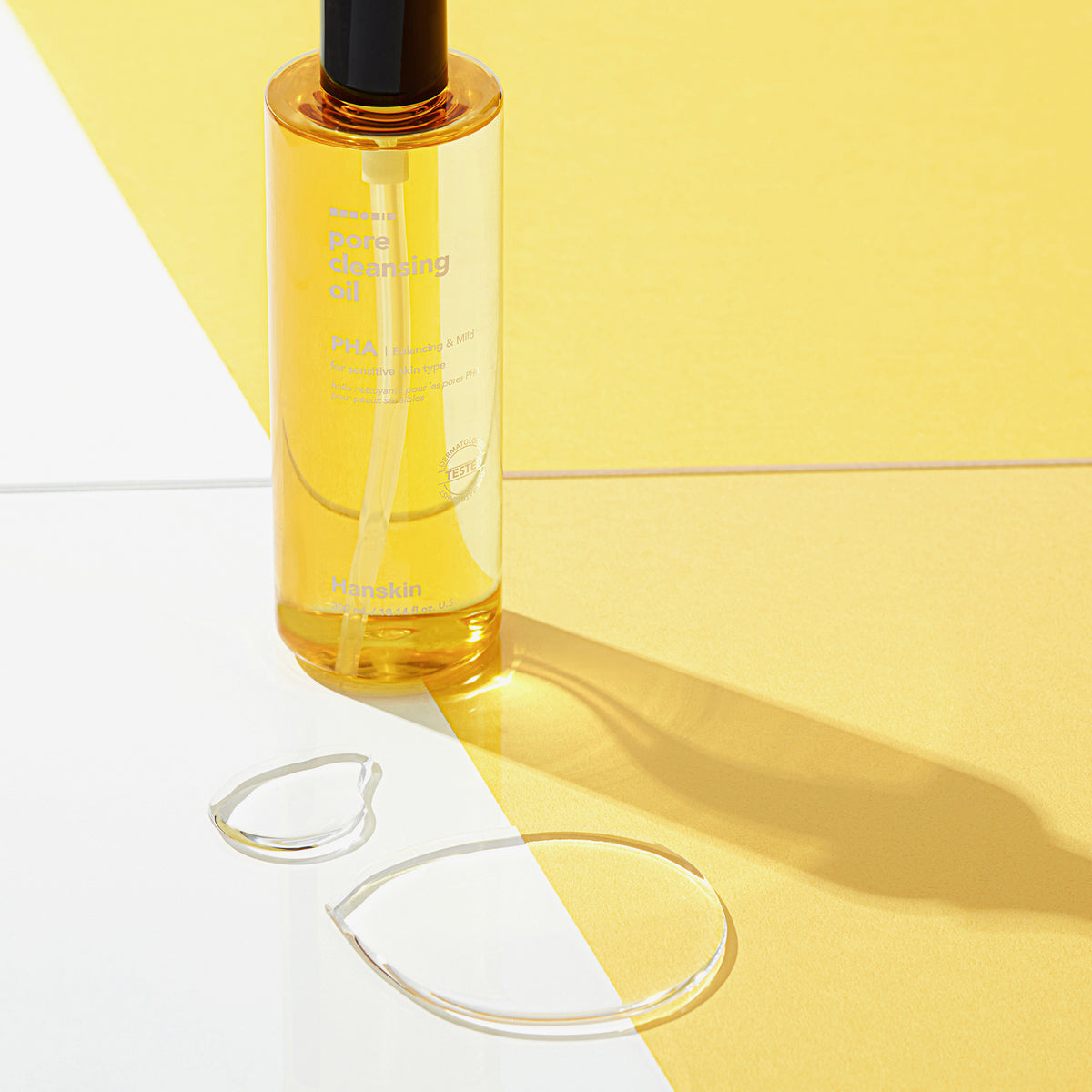 Eine Flasche Pore Cleansing Oil PHA mit zwei Tropfen Öl auf gelb-weißem Hintergrund.