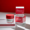 Ein Tiegel und zwei Schachteln Vitamin A Glow Retinol Cream werden auf einer weißen Fläche vor einem roten Hintergrund präsentiert.