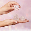 Eine Hand hält einen Deckel und zwei  Fix Potion-Tiegel  gefüllt mit klarem Gel vor rosa Bokeh-Hintergrund.
