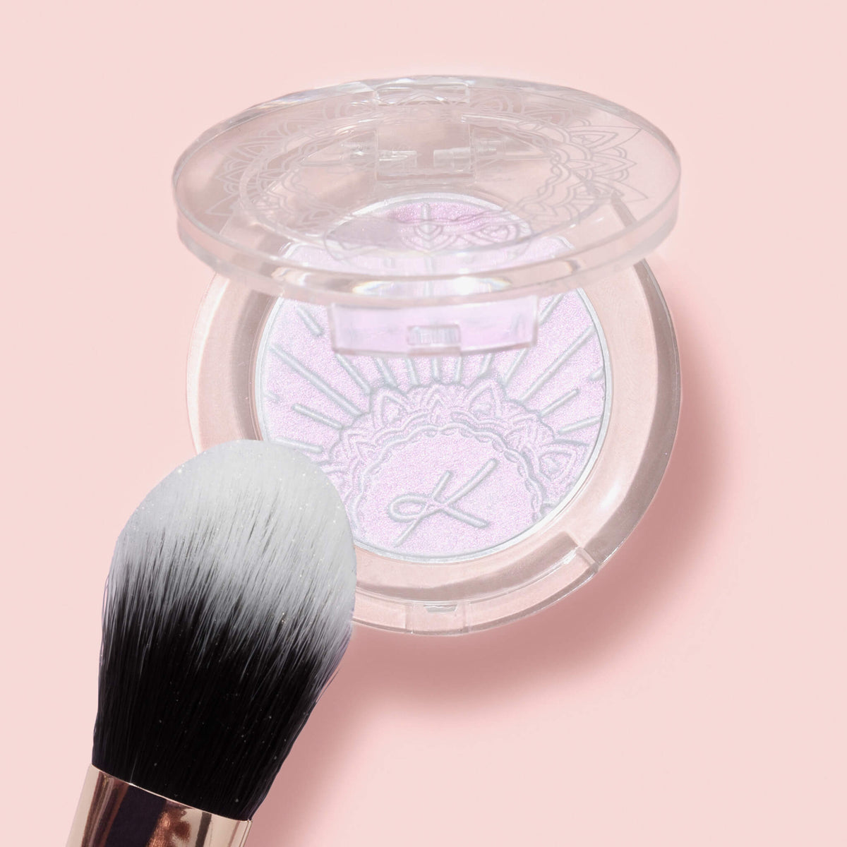 Der K10: Highlighter Brush liegt neben einer offenen Dose mit schimmerndem gepresstem Puder auf pastellrosa Hintergrund.