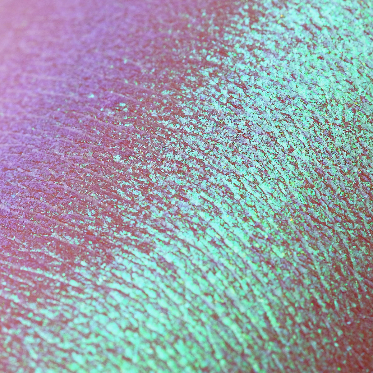 Nahaufnahme der Textur der Pressed Opal Multichrome Eyeshadow Birdsong, die rosa und türkis auf der Haut schimmert.