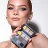Eine Frau mit auffälligem Lidstrich hält die LH Cosmetics Aim Higher Palette mit verschiedenen Farbtönen vor ihr Gesicht und lächelt.