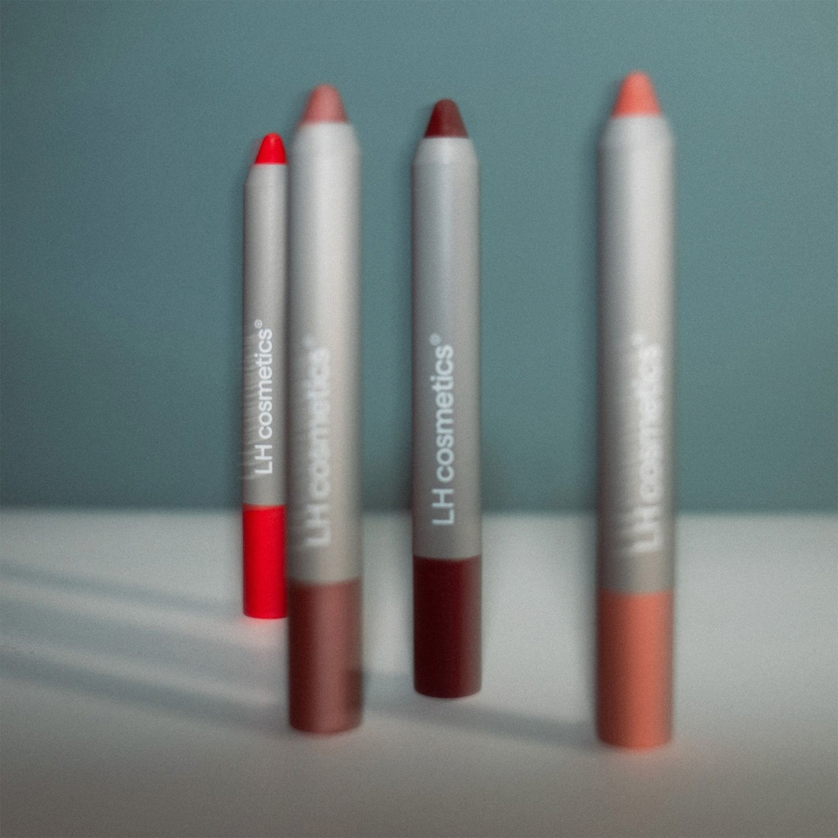 Vier LH Cosmetics Biggie Lippenstifte in verschiedenen Farbtönen stehen aufrecht auf einer weißen Fläche mit blauem Hintergrund.