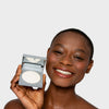Eine lächelnde Person hält vor einem schlichten Hintergrund ein geöffnetes LH Cosmetics Blotting Powder mit einem Spiegel.