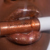 Nahaufnahme orange-gold glitzernder Lippen, die eine Glazed-Lipgloss-Tube in der Farbe Melt von LH Cosmetics zwischen den Zähnen halten.