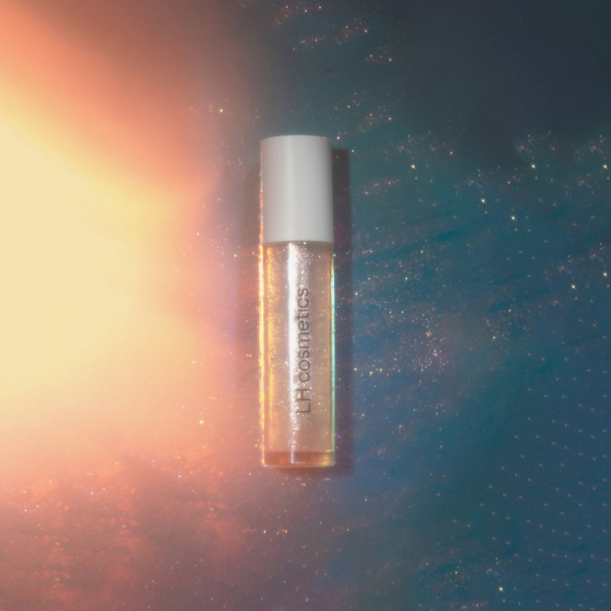 Transparente Tube Glazed Yum von LH Cosmetics mit weißem Deckel und glitzerndem Inhalt liegt auf blau und gelb leuchtender Oberfläche.