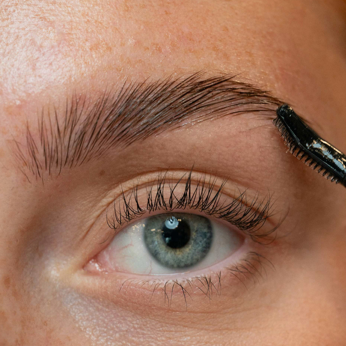 Eine Nahaufnahme eines blauen Auges, während jemand seine Augenbrauen mit dem The Brow Wax von LH Cosmetics pflegt.