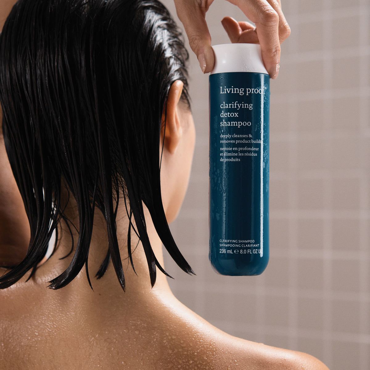 Eine Person mit nassen Haaren hält unter der Dusche eine Flasche Living Proof Clarifying Detox Shampoo.
