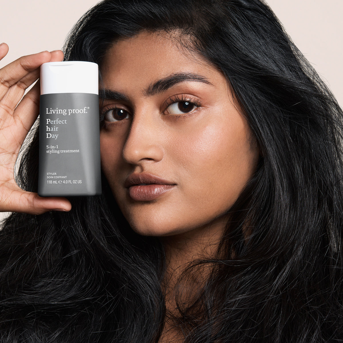 Frau mit langem schwarzen Haar hält ein Living Proof Perfect Hair Day 5-in-1 Styling Treatment neben ihr Gesicht.