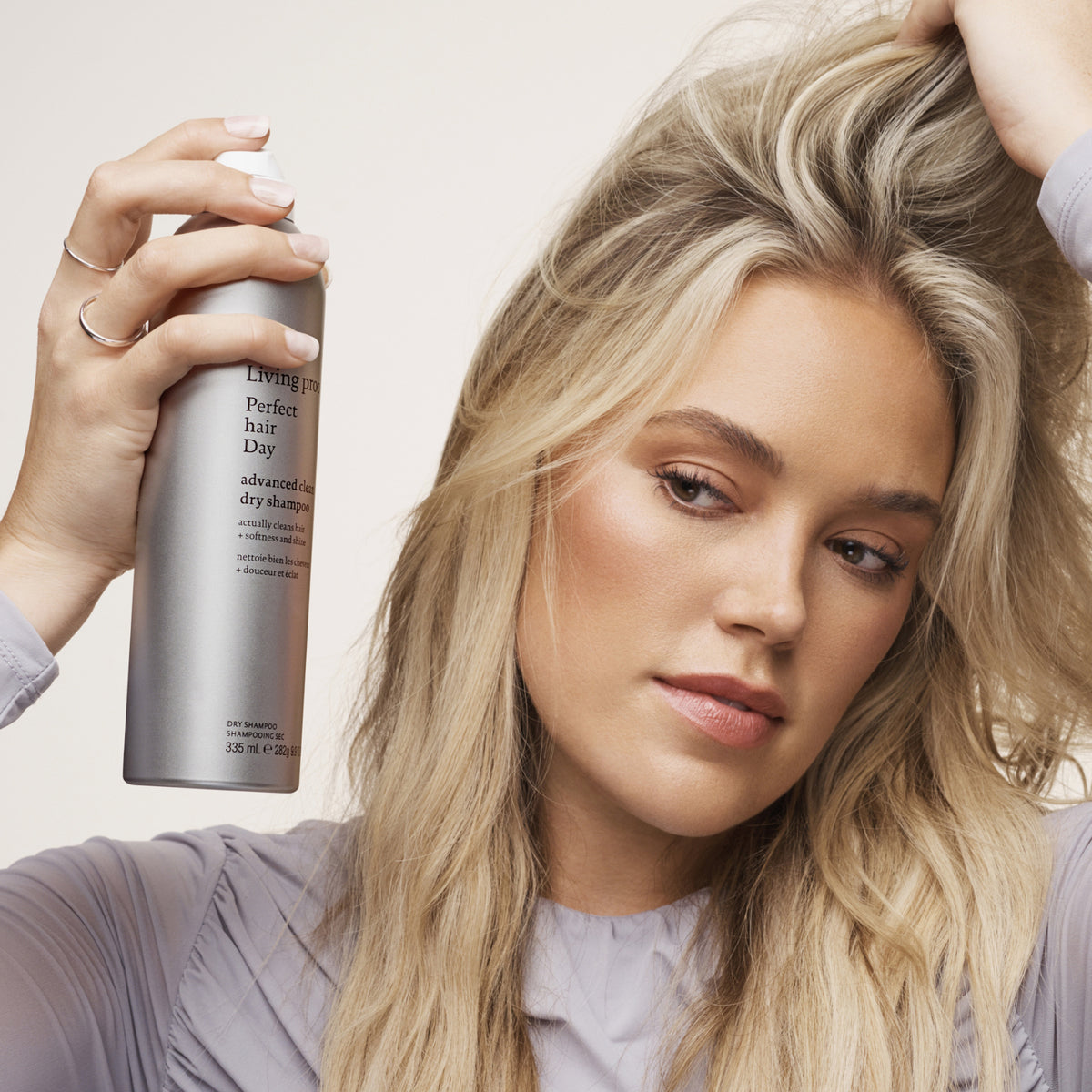 Eine Frau sprüht Living Proof Perfect Hair Day Advanced Clean Dry Shampoo auf ihr blondes Haar, während sie es mit der Hand anhebt.