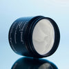 Geöffneter Tiegel Style Lab Amp Texture Volumizer von Living Proof mit weißem Produkt, präsentiert auf einer glänzenden hellblauen Oberfläche.