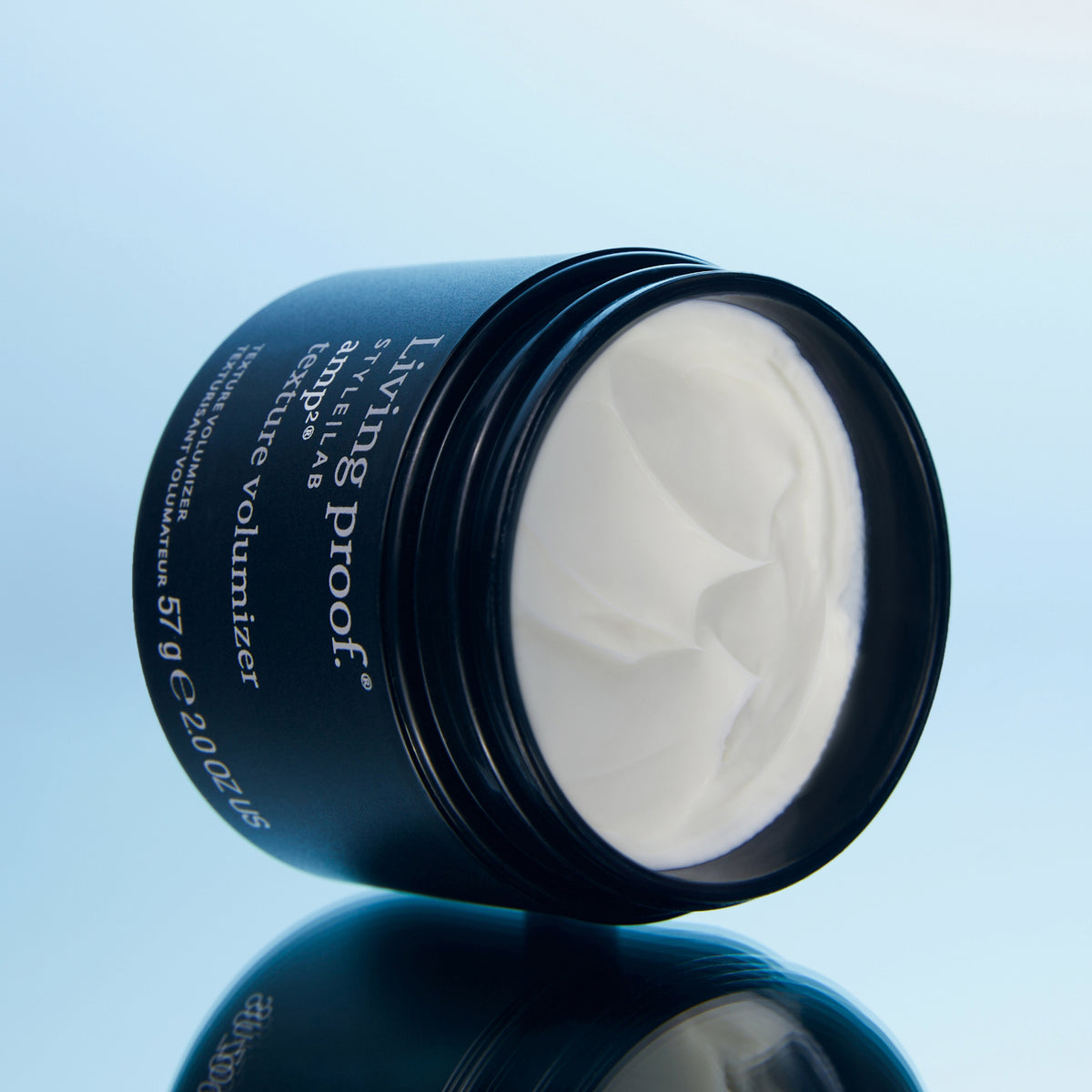 Geöffneter Tiegel Style Lab Amp Texture Volumizer von Living Proof mit weißem Produkt, präsentiert auf einer glänzenden hellblauen Oberfläche.