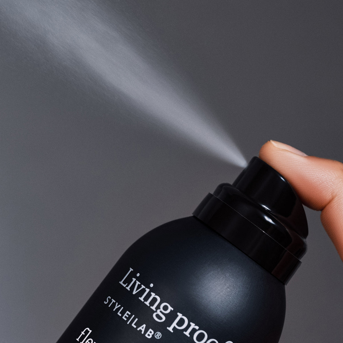 Eine Hand sprüht Living Proof Style Lab Flex Haarspray vor einem grauen Hintergrund.
