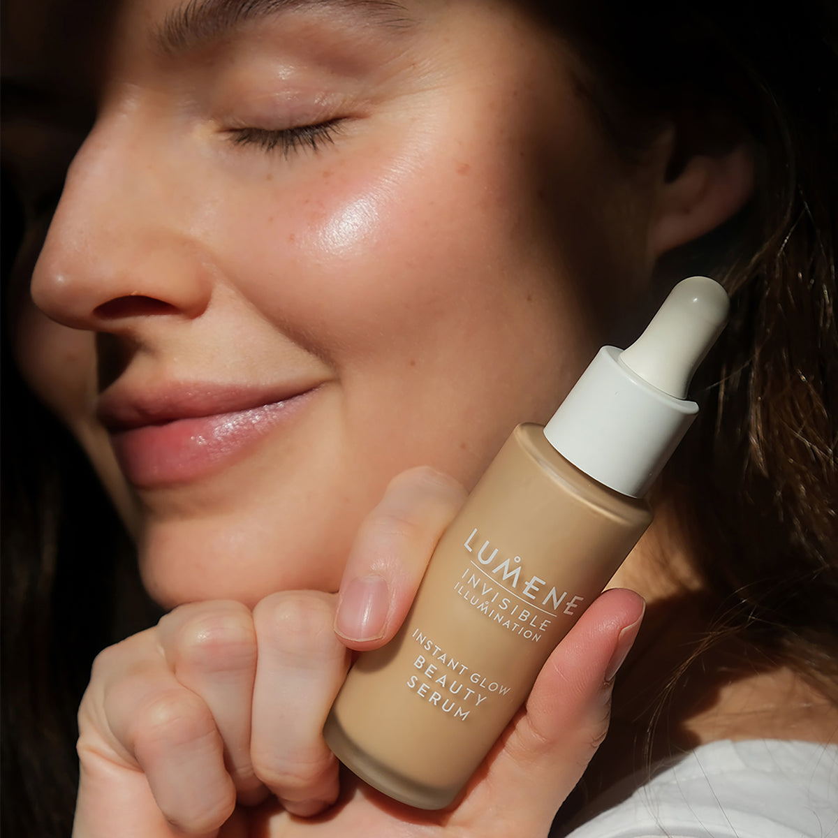 Eine lächelnde Frau mit strahlender Haut hält eine Flasche Lumene INVISIBLE ILLUMINATION Glow Beauty Serum in der Nähe ihres Gesichts.