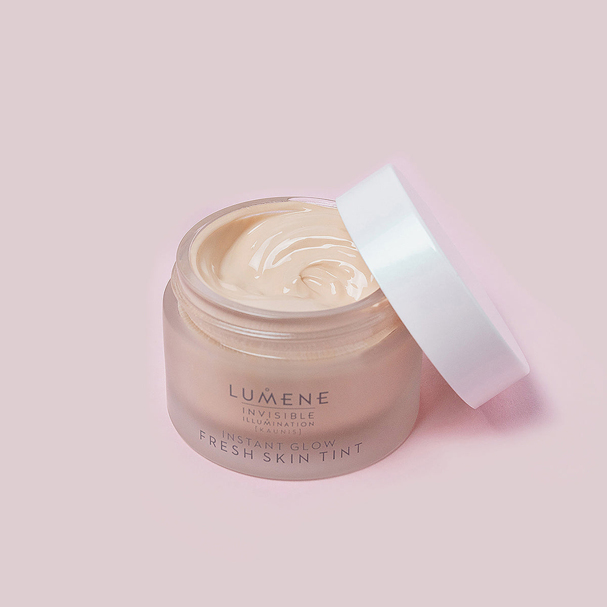 Eine geöffnete Dose Lumene INVISIBLE ILLUMINATION Instant Glow Fresh Skin Tint mit weißem Deckel ist vor einem hellrosa Hintergrund abgebildet.