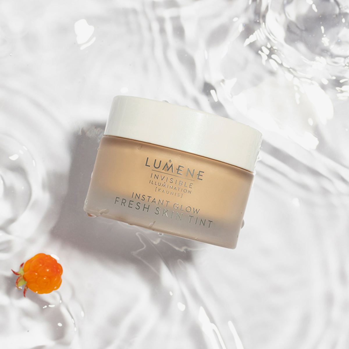 Ein Tiegel Lumene INVISIBLE ILLUMINATION Instant Glow Fresh Skin Tint auf einer Wasseroberfläche mit einer orangefarbenen Beere daneben.