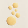 Mehrere beige Tropfen Lumene NORDIC-C [VALO] Glow Boost Eye Serum auf einer leicht strukturierten hellen Oberfläche.
