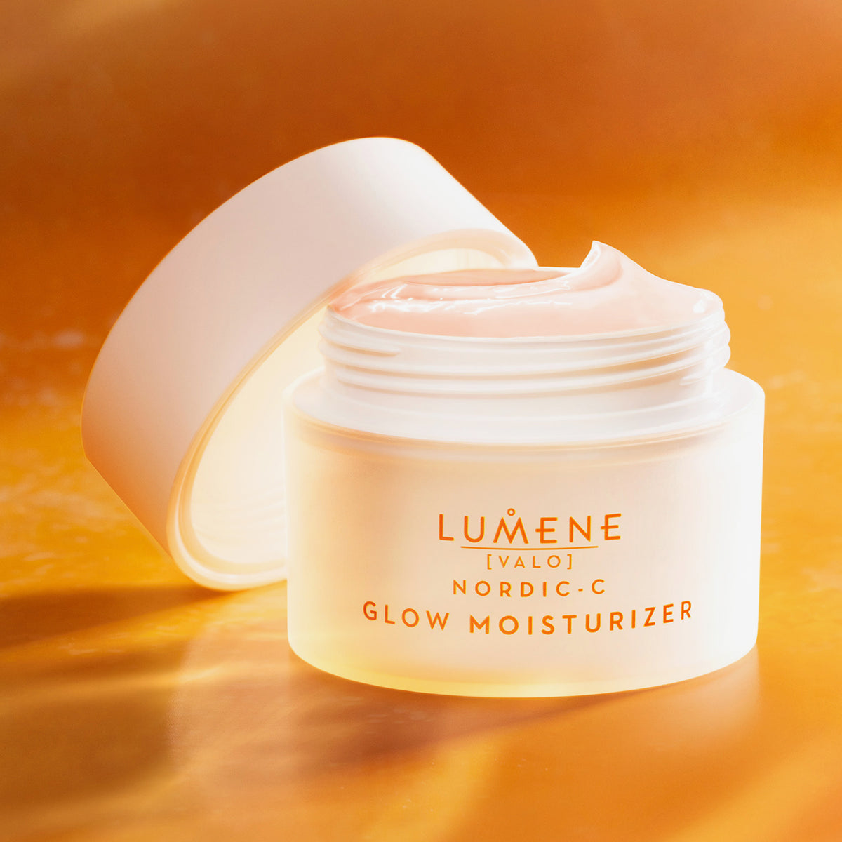 Ein geöffneter weißer Tiegel Lumene NORDIC-C [VALO] Glow Moisturizer ist auf einem orangefarbenen Hintergrund abgebildet.