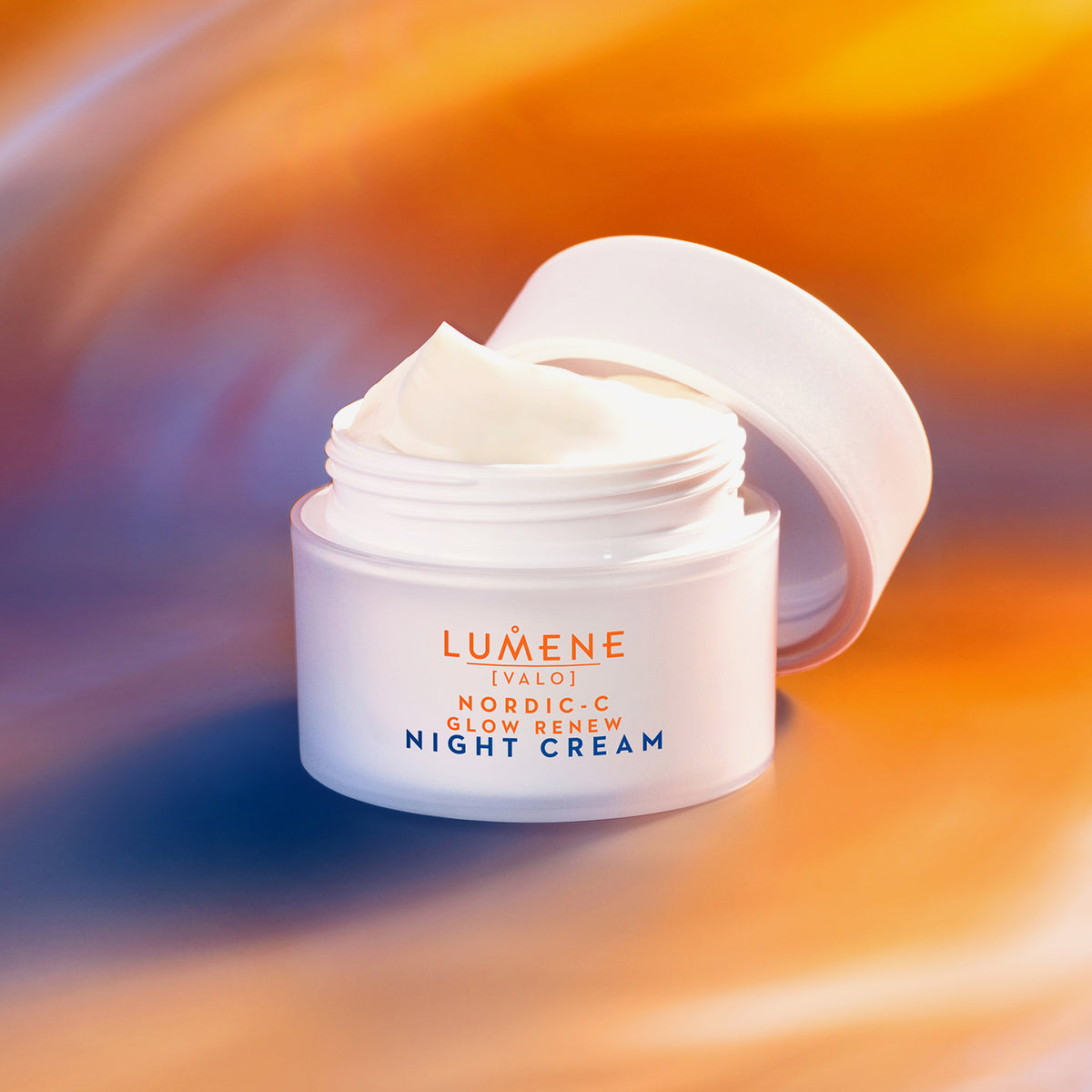 Ein geöffneter weißer Tiegel der Lumene Nordic-C [VALO] Glow Renew Night Cream vor einem leuchtenden, orangefarbenen und blauen, verschwommenen Hintergrund.