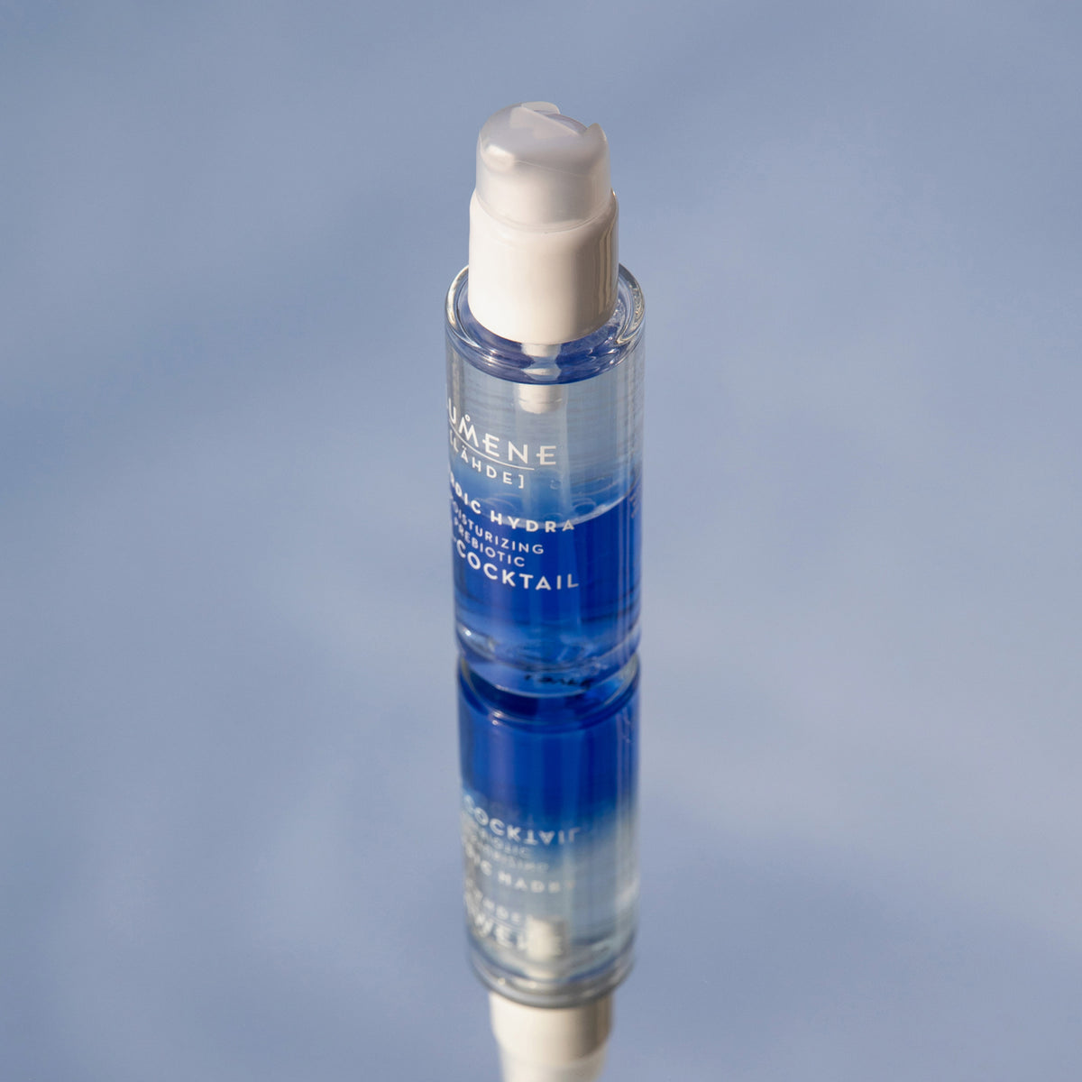 Eine Flasche Lumene NORDIC HYDRA [LAHDE] Moisturizing Prebiotic Oil-Cocktail, gefüllt mit blauer und transparenter Flüssigkeit, spiegelt sich auf einer glatten, hellblauen Oberfläche.