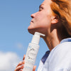 Rothaarige Frau hält eine Flasche Lumene NORDIC HYDRA [LAHDE] Oat Milk Oil Cleanser mit geschlossenen Augen und nach oben geneigtem Kopf vor einem Hintergrund mit blauem Himmel.