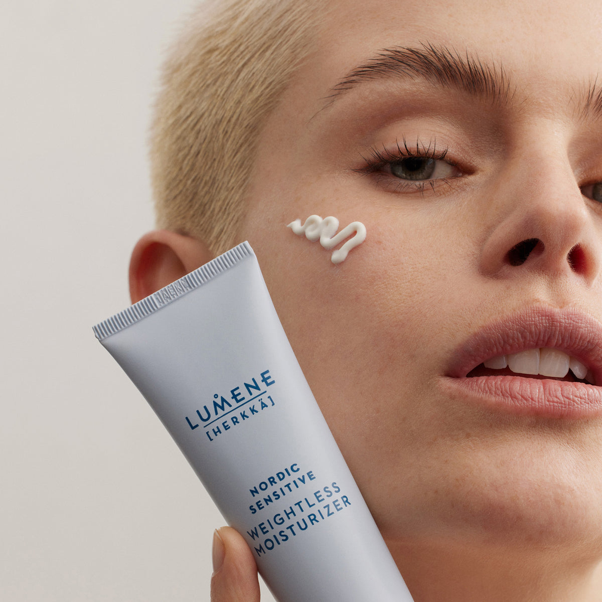 Eine Person mit kurzen blondierten Haaren trägt Lumene Nordic Sensitive [HERKKÄ] Weightless Moisturizer auf ihre Wange auf.