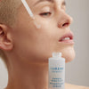 Person mit kurzem blondierten Haar trägt Lumene Nordic Sensitive [HERKKÄ] Weightless Serum mit einer Pipette auf die Wange auf.