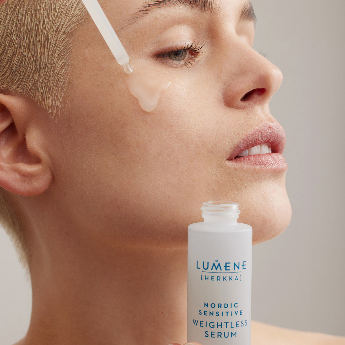 Person mit kurzem blondierten Haar trägt Lumene Nordic Sensitive [HERKKÄ] Weightless Serum mit einer Pipette auf die Wange auf.