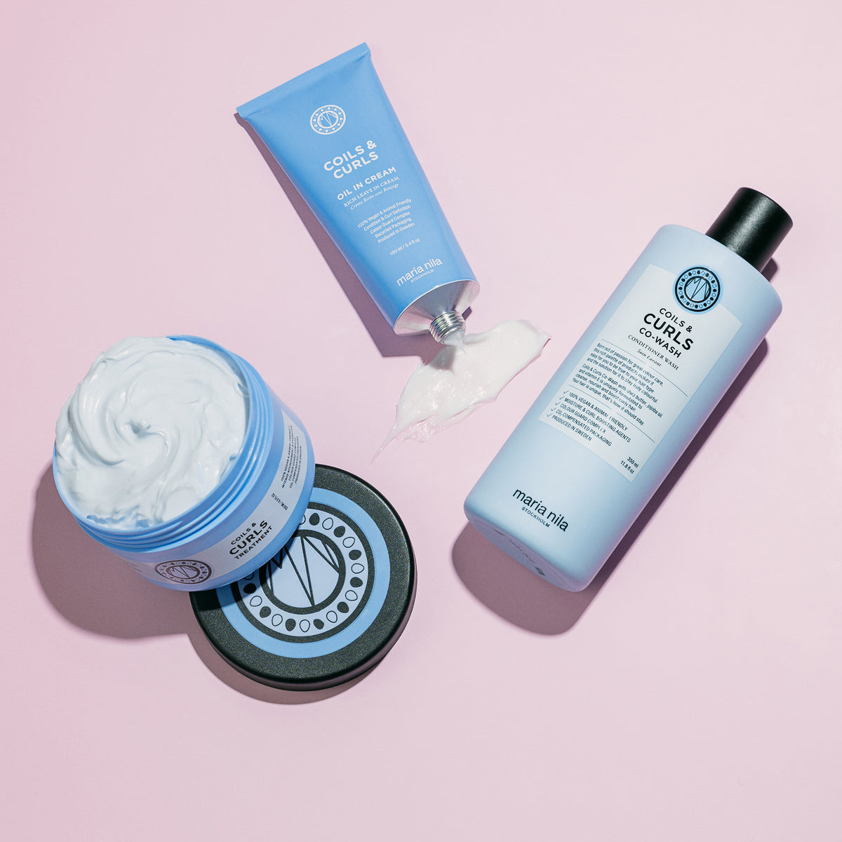 Die maria nila Coils & Curls Oil-In-Cream in hellblauer Verpackung neben weiteren Coils & Curls Produkten sind auf einem rosa Hintergrund abgebildet.